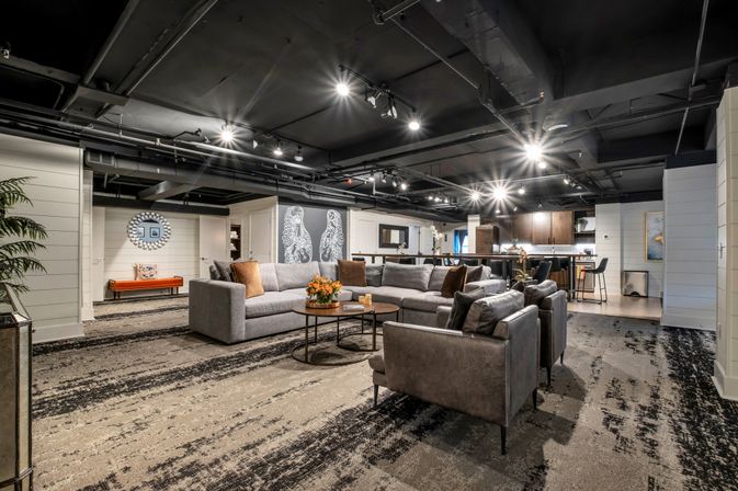 Spacious modern finished basement lounge — cozy gray sectional and leather armchairs around round coffee tables, open kitchen with bar stools, industrial black ceiling with track lighting, patterned carpet and decorative wall art.