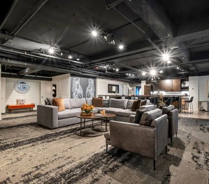 Spacious modern finished basement lounge — cozy gray sectional and leather armchairs around round coffee tables, open kitchen with bar stools, industrial black ceiling with track lighting, patterned carpet and decorative wall art.