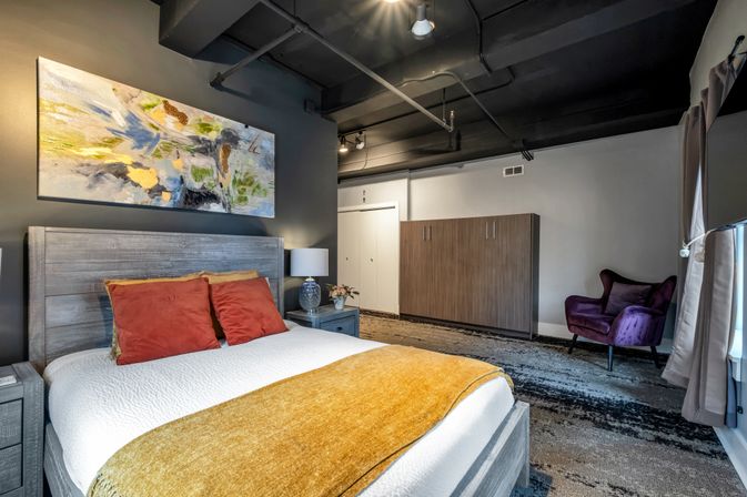 Stylish urban loft bedroom with exposed black industrial ceiling, gray wood bed with rust‑orange pillows and mustard throw, abstract wall art and purple accent chair