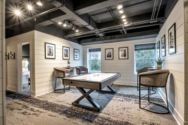 Inviting modern game room in an apartment clubhouse with an air hockey table, plush swivel chairs, shiplap walls, framed photos, large windows and exposed industrial ceiling