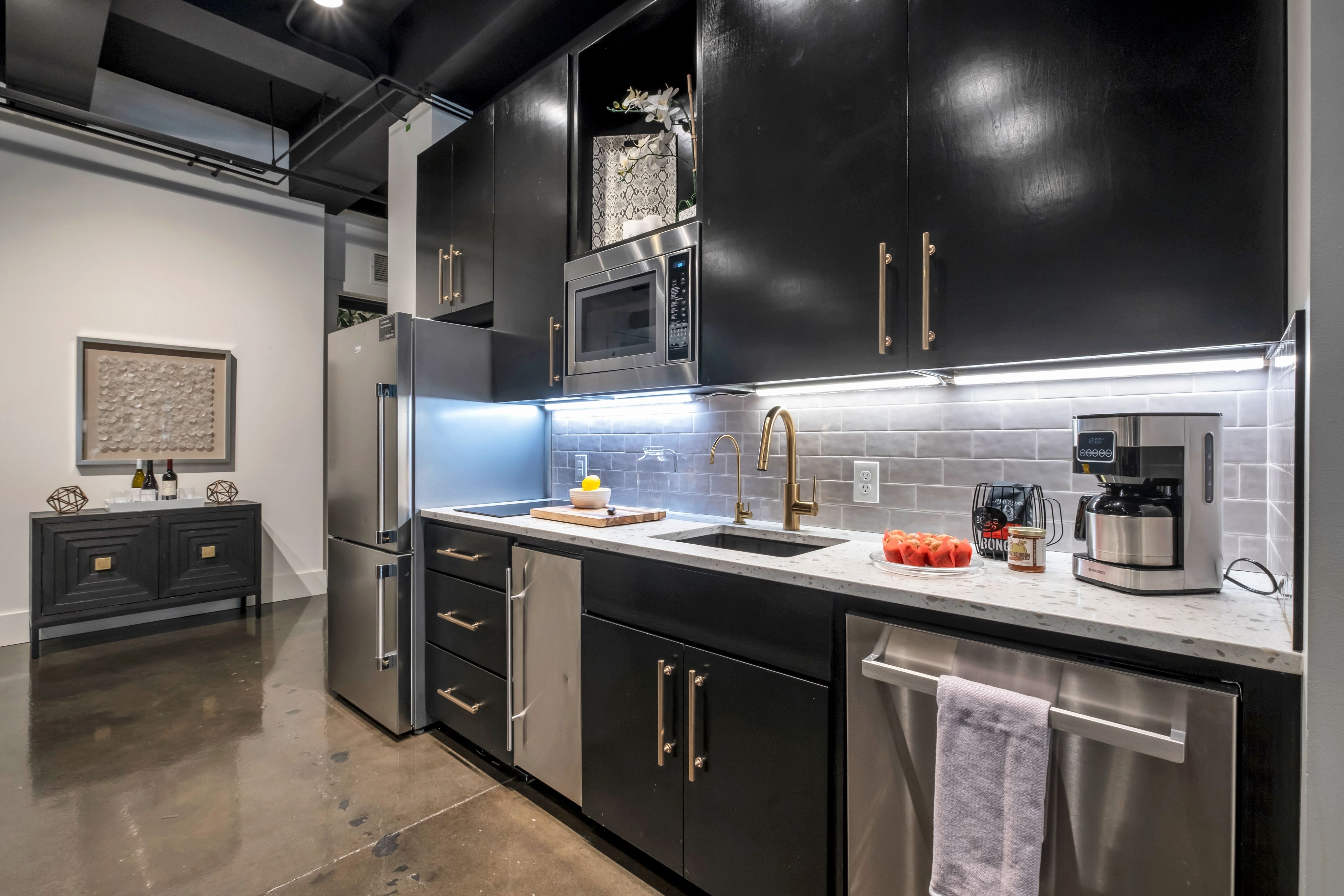 Modern kitchen with glossy black cabinets, stainless steel fridge, microwave and dishwasher, quartz countertop, gold faucet, gray subway tile backsplash, under-cabinet LED lights, coffee maker and bowl of strawberries on the counter, polished concrete floor.