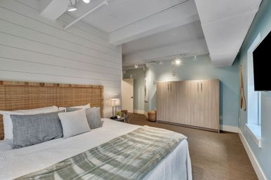 Coastal-chic bedroom with woven rattan headboard, white shiplap wall, king bed with gray pillows and plaid throw, soft blue walls, wood storage cabinet and track lighting.