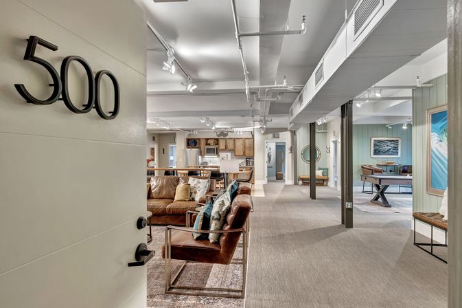 Door labeled 500 opening into a cozy, modern open-concept lounge and kitchen with brown leather seating, accent pillows, exposed industrial ceiling, and a game table