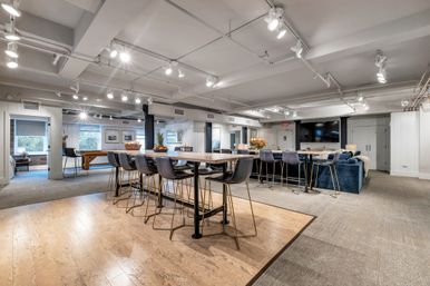 Open modern communal lounge with long high-top tables and brass-legged bar stools, blue velvet sectional facing a large wall-mounted TV, pool table near windows, warm wood and carpet flooring, bright track lighting and decorative plants