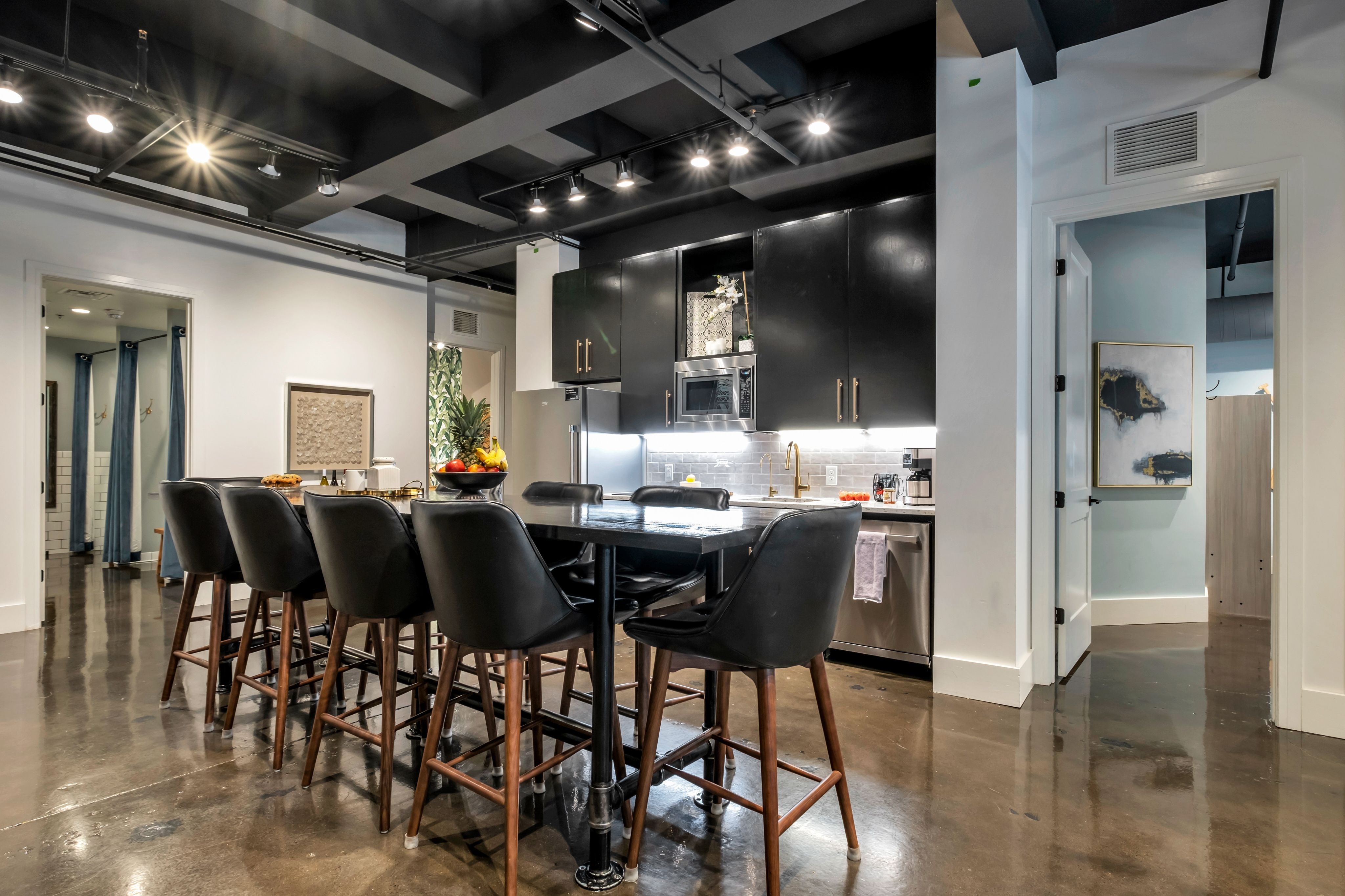 Sleek modern open-concept kitchen with matte black cabinets, stainless appliances, long high-top island with six leather bar stools, track lighting and polished concrete floor