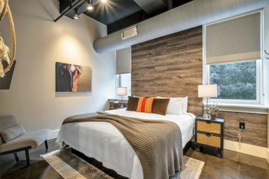 Cozy modern industrial bedroom with wood accent wall, white bed topped by a brown knit throw and striped pillow, rattan nightstand with lamp, exposed ductwork, and horse painting