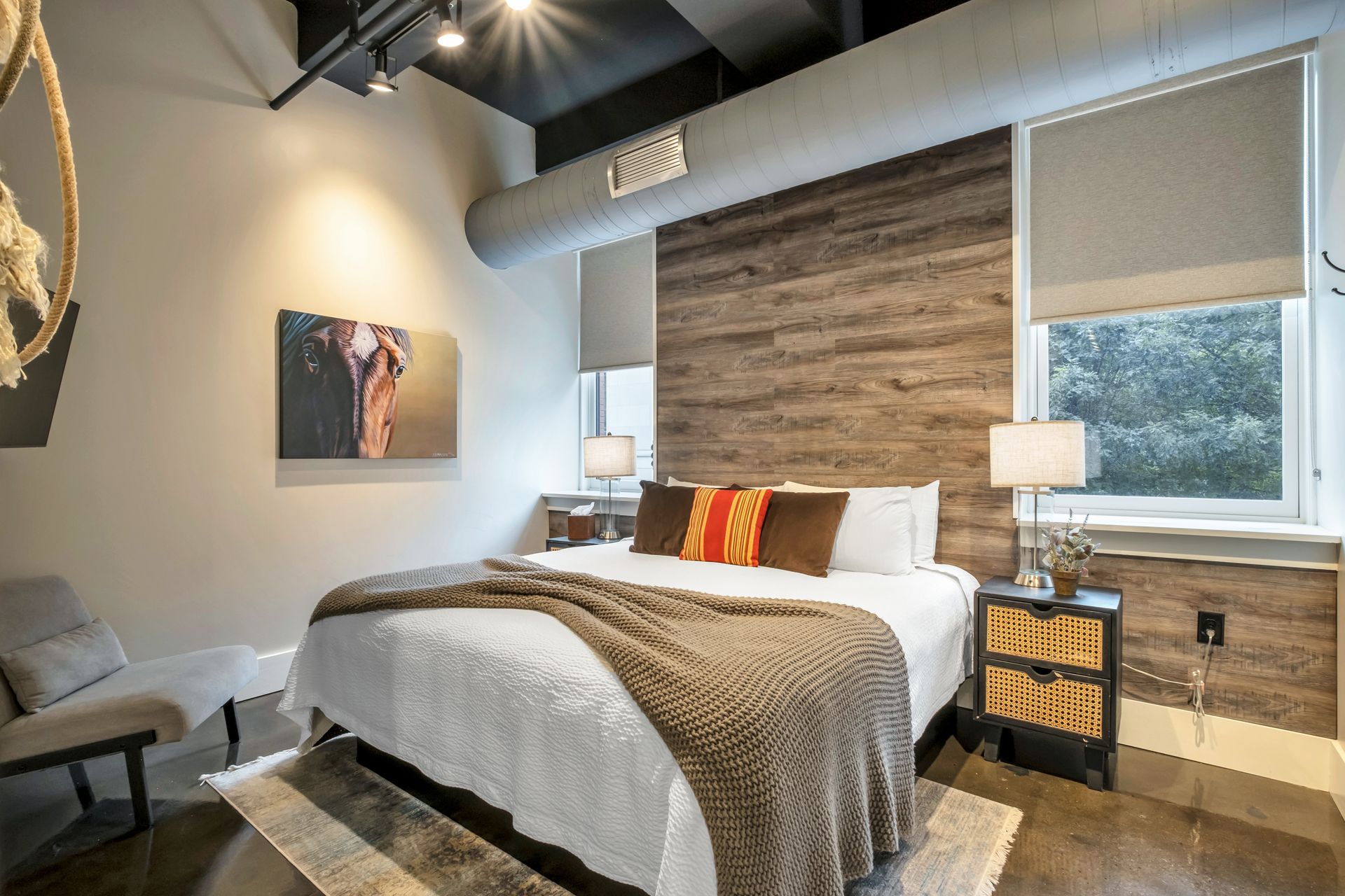 Cozy modern industrial bedroom with wood accent wall, white bed topped by a brown knit throw and striped pillow, rattan nightstand with lamp, exposed ductwork, and horse painting