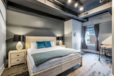 Cozy modern industrial urban loft bedroom with light wood king bed, teal accent pillows, textured gray walls, bedside lamps and a city window.