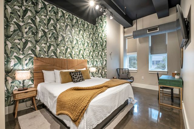 Modern urban loft bedroom with tropical leaf wallpaper, woven rattan headboard, king bed with white linens and mustard knit throw, bedside lamp, wicker chair, green desk, exposed ductwork and polished concrete floor