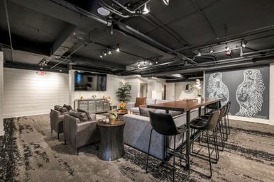 Stylish community lounge with sectional sofa, leather armchairs, high wooden table with stools, wall-mounted TV, and large chalk-style winged guitar mural.