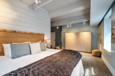 Coastal-style bedroom with rattan headboard and white shiplap wall, king bed dressed in gray pillows and a plush brown throw, bedside lamp, light-wood wardrobe, woven pouf and soft track lighting