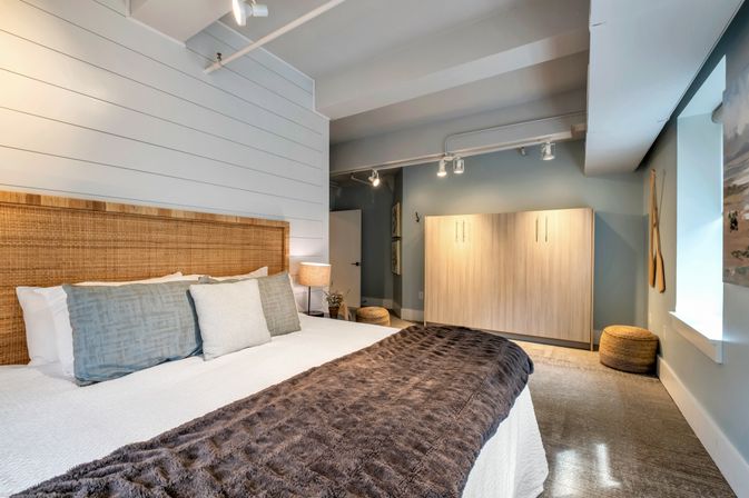 Coastal-style bedroom with rattan headboard and white shiplap wall, king bed dressed in gray pillows and a plush brown throw, bedside lamp, light-wood wardrobe, woven pouf and soft track lighting
