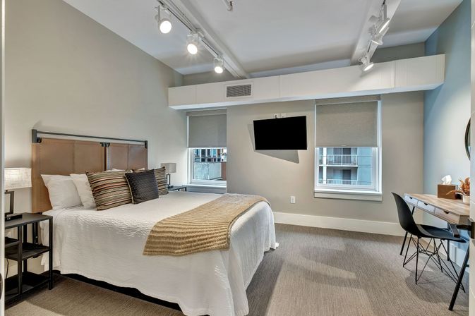 Cozy modern urban apartment bedroom with large bed and leather headboard, white linens and beige throw, striped accent pillows, track lighting, wall-mounted TV, work desk and two windows with roller shades overlooking nearby city buildings