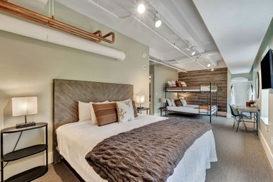 Modern urban loft-style bedroom with king bed, metal bunk beds, wood accent wall, exposed copper pipes and track lighting, neutral cozy bedding and small desk workspace