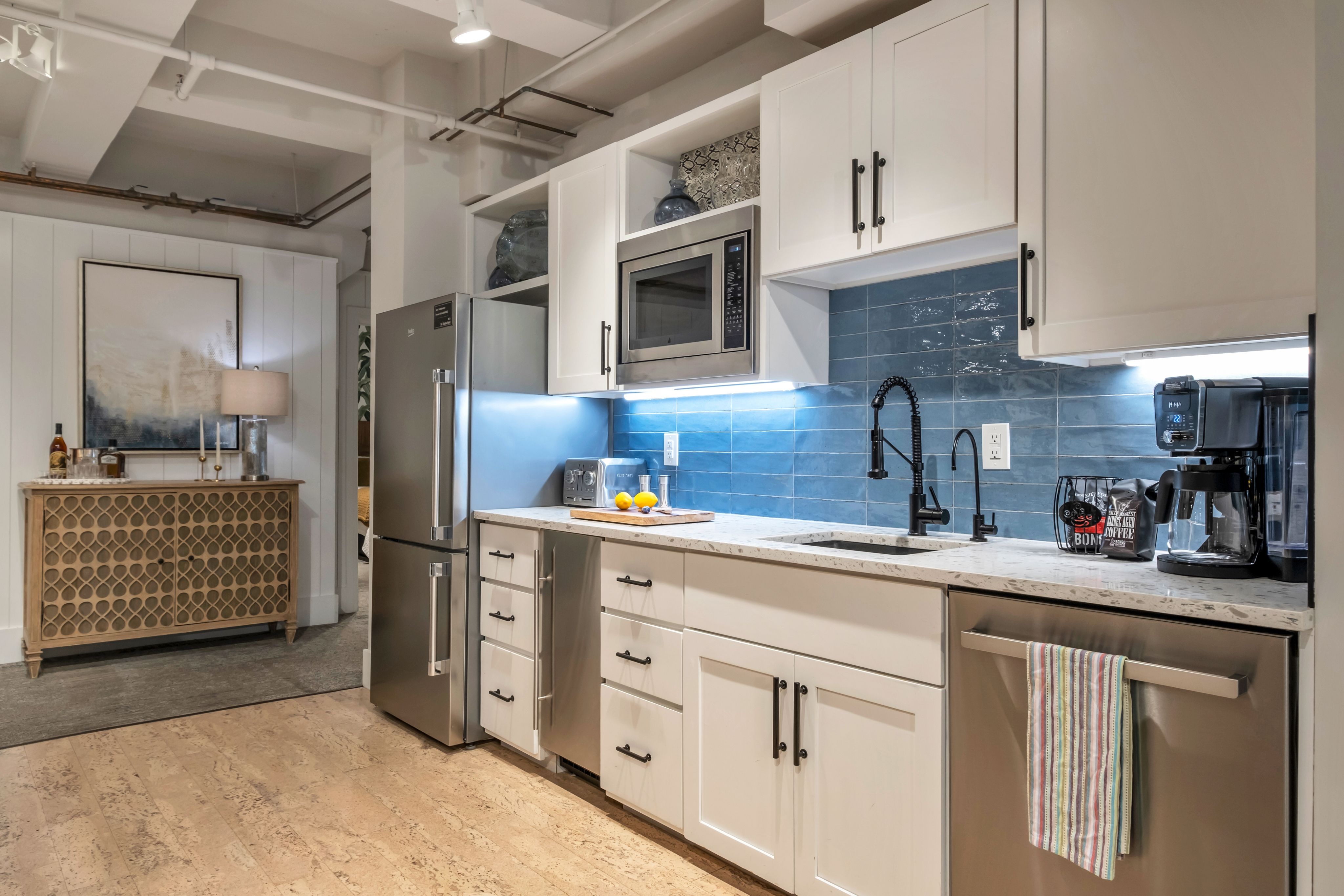 Bright modern kitchen interior with white shaker cabinets, blue subway-tile backsplash, stainless-steel fridge, microwave and dishwasher, black gooseneck faucet, quartz countertop with lemons and coffee maker, light wood floor