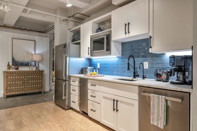 Bright modern kitchen interior with white shaker cabinets, blue subway-tile backsplash, stainless-steel fridge, microwave and dishwasher, black gooseneck faucet, quartz countertop with lemons and coffee maker, light wood floor