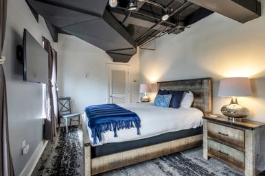 Cozy industrial-style loft bedroom with reclaimed-wood bed and nightstand, white bedding and navy blue throw, decorative pillows, metallic bedside lamps, wall-mounted TV, gray curtains and exposed black ceiling beams.