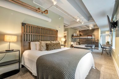 Cozy modern loft bedroom with wooden king headboard, white linens and gray knit throw, metal bunk beds in the background, small desk and wall-mounted TV, exposed ceiling pipes and warm bedside lamps