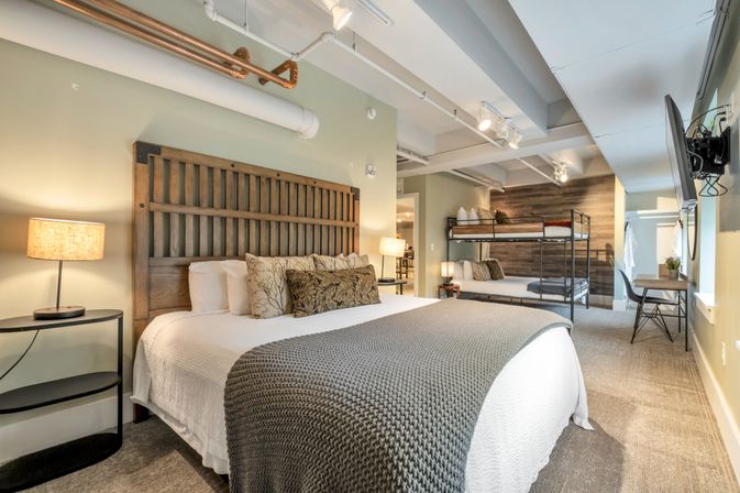 Cozy modern loft bedroom with wooden king headboard, white linens and gray knit throw, metal bunk beds in the background, small desk and wall-mounted TV, exposed ceiling pipes and warm bedside lamps