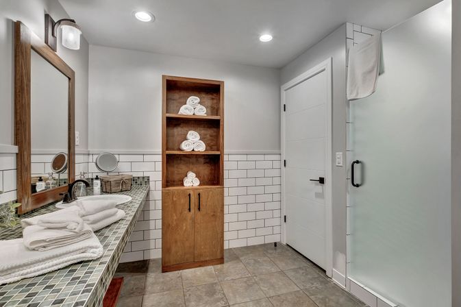 Modern spa-ready residential bathroom with white subway tile, wooden open shelving of rolled towels, frosted glass shower, mosaic-tiled vanity, sink and mirror.
