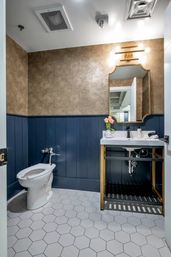 Chic modern powder room with navy wainscoting, textured gold wallpaper, white hexagon tile floor, white toilet, gold-framed mirror and sink with brass sconce and tulips