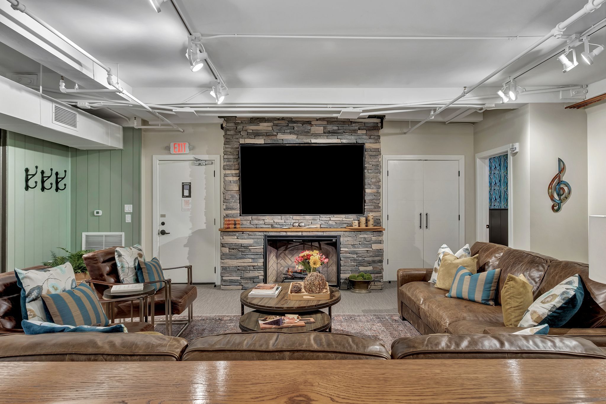 Cozy modern rustic basement lounge with stacked stone fireplace and large mounted flat-screen TV, brown leather sofas with colorful throw pillows, round wooden coffee tables with floral centerpiece, exposed white ceiling pipes and track lighting, green shiplap accent wall with coat hooks.