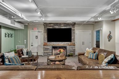 Cozy modern rustic basement lounge with stacked stone fireplace and large mounted flat-screen TV, brown leather sofas with colorful throw pillows, round wooden coffee tables with floral centerpiece, exposed white ceiling pipes and track lighting, green shiplap accent wall with coat hooks.