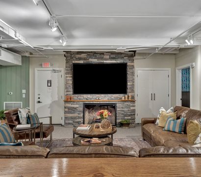 Cozy modern rustic basement lounge with stacked stone fireplace and large mounted flat-screen TV, brown leather sofas with colorful throw pillows, round wooden coffee tables with floral centerpiece, exposed white ceiling pipes and track lighting, green shiplap accent wall with coat hooks.