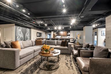 Modern open-concept basement living room with a large gray sectional, leather armchairs, round wood coffee table with orange flowers, exposed black industrial ceiling and track lighting, and a kitchen island in the background — cozy urban-chic.