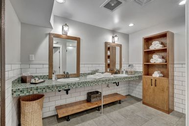 Spa-style residential bathroom with double sinks on green mosaic tile countertop, two wood-framed mirrors and sconces, white subway tile walls, open wooden shelving with rolled white towels, woven laundry basket and wood bench on gray tiled floor.