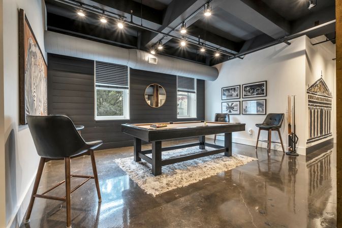 Loft-style urban game room with a black pool table on a shag rug, leather bar stools, polished concrete floor, exposed ductwork, round mirror and framed wall art.