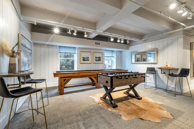 Stylish modern game room interior with a foosball table on a cowhide rug, long wooden shuffleboard, white shiplap walls, two windows, track lighting and black bar stools at small wall-mounted tables.