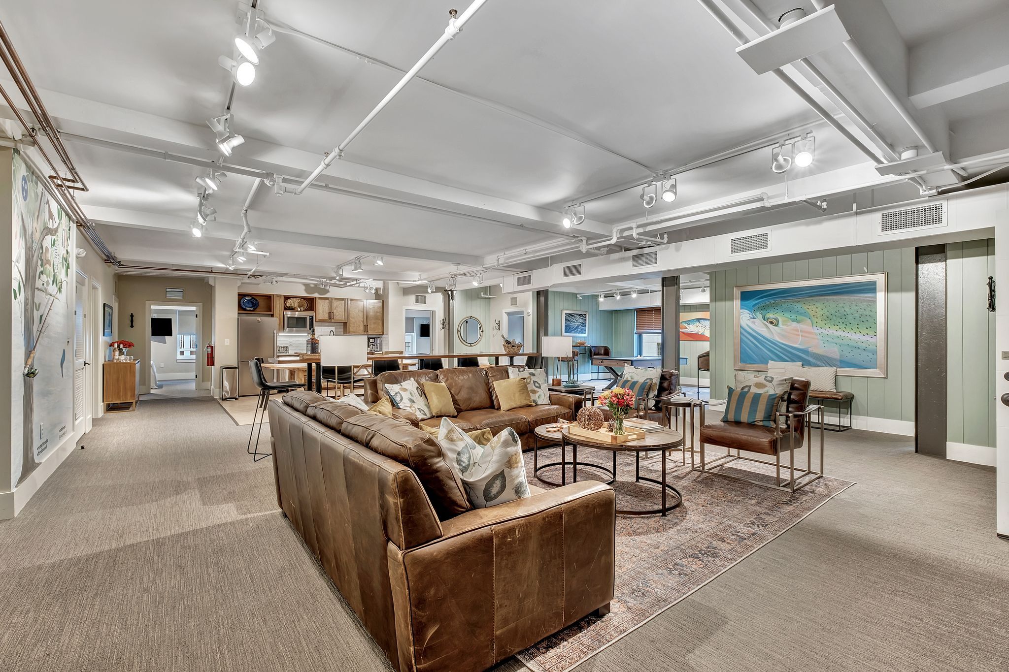 Spacious finished basement lounge with brown leather sofas, round coffee tables on a patterned rug, kitchen bar seating, modern track lighting and framed fish artwork for a relaxed coastal vibe.