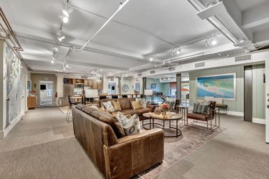 Spacious finished basement lounge with brown leather sofas, round coffee tables on a patterned rug, kitchen bar seating, modern track lighting and framed fish artwork for a relaxed coastal vibe.
