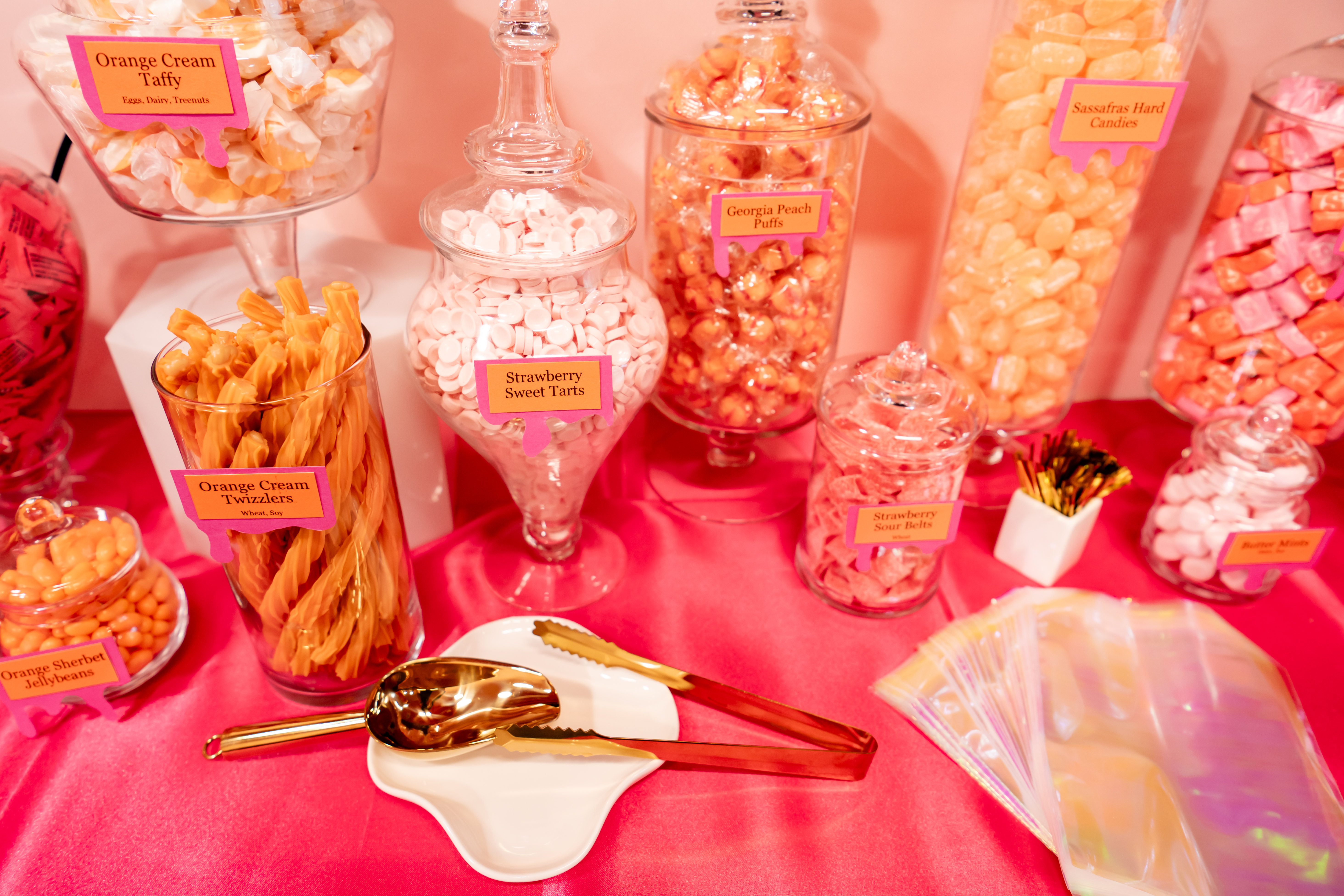 Playful pink candy buffet with glass apothecary jars and clear containers of orange and pink sweets—orange cream twizzlers, strawberry sweet tarts and peach puffs—gold tongs and iridescent treat bags on a hot-pink tablecloth.