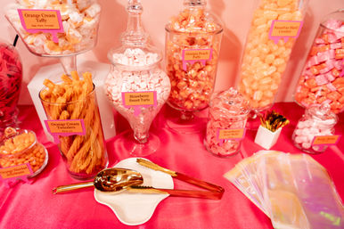 Playful pink candy buffet with glass apothecary jars and clear containers of orange and pink sweets—orange cream twizzlers, strawberry sweet tarts and peach puffs—gold tongs and iridescent treat bags on a hot-pink tablecloth.