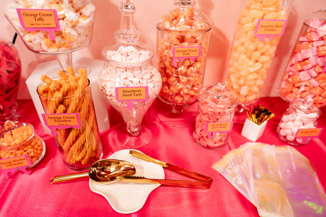 Playful pink candy buffet with glass apothecary jars and clear containers of orange and pink sweets—orange cream twizzlers, strawberry sweet tarts and peach puffs—gold tongs and iridescent treat bags on a hot-pink tablecloth.