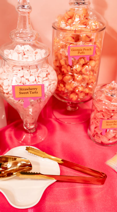 Playful pink candy buffet with glass apothecary jars and clear containers of orange and pink sweets—orange cream twizzlers, strawberry sweet tarts and peach puffs—gold tongs and iridescent treat bags on a hot-pink tablecloth.