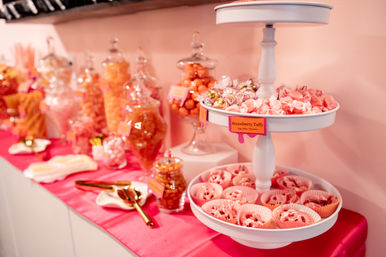 Pink candy buffet with glass apothecary jars and a white tiered tray filled with strawberry taffy and pink confections on a bright pink tablecloth