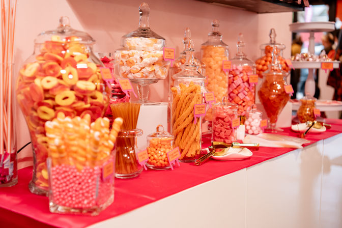 Colorful pink-and-orange candy buffet with assorted sweets in glass jars and scoops on a red table runner at an event