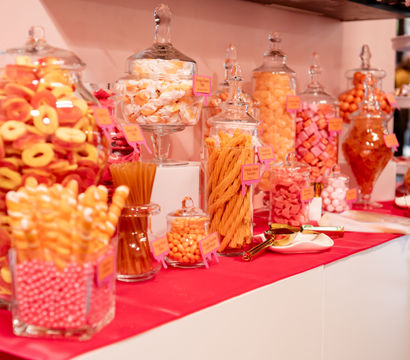 Colorful pink-and-orange candy buffet with assorted sweets in glass jars and scoops on a red table runner at an event