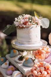Semi-naked single-tier wedding cake topped with pink roses, baby’s breath and eucalyptus on a glass stand, surrounded by pastel chocolate-covered strawberries and bowls of pink popcorn on an outdoor garden dessert table.