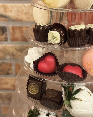 Tiered dessert display of gourmet chocolates and macarons: red heart-shaped chocolates, dark cups with white drizzle, gold-stamped chocolate squares, white chocolate–dipped strawberries and pastel macarons against a brick wall.