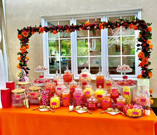 Orange-themed candy buffet with glass jars of pink, yellow and orange sweets arranged on an orange tablecloth beneath a garland arch of orange roses, tiered dessert stands and a white-framed window backdrop.