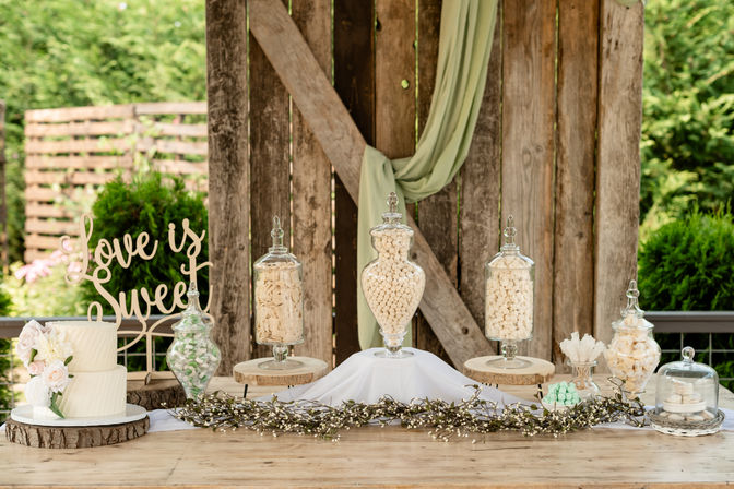 Rustic outdoor wedding candy table with barn-style wooden backdrop and greenery, white tiered cake, 'Love is Sweet' sign, and glass apothecary jars on wood slices filled with white and pastel candies