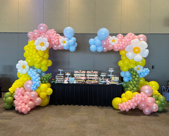 Playful pastel balloon arch with daisy-shaped balloons framing a dessert table in an indoor event hall — pink, yellow, blue party decor