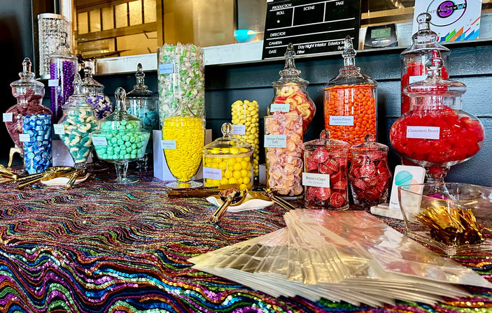 Colorful candy buffet with glass apothecary jars of assorted candies (red gummies, orange-coated candies, peach taffy, yellow lemon candies, mint greens) arranged on a shimmering multicolored sequin tablecloth at an event