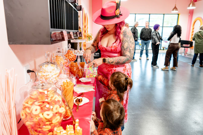 Stylish woman in a pink hat and floral dress with tattooed arms fills a treat bag at a bright pink candy buffet of orange and pink sweets while two children watch in a modern indoor party space.