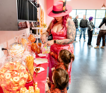 Stylish woman in a pink hat and floral dress with tattooed arms fills a treat bag at a bright pink candy buffet of orange and pink sweets while two children watch in a modern indoor party space.