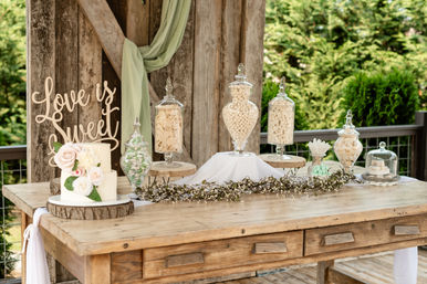 Rustic outdoor wedding dessert table on a wooden table with a "Love is Sweet" sign, two-tier white cake with blush roses, glass apothecary jars filled with white candies, wood slice stands and a delicate greenery garland against a wood backdrop and garden foliage.